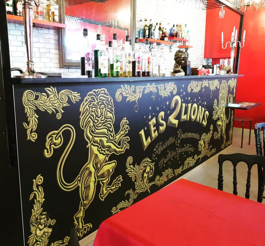Restaurant cabaret Les 2 Lions Téléphone : 06 15 70 76 23 Mur décoré avec un lion doré et le texte "LES 2 LIONS" dans un bar. Tables rouges.